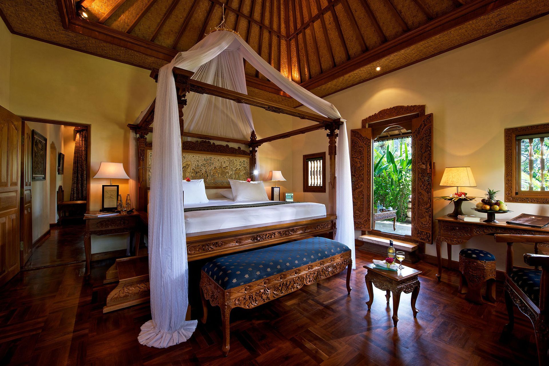 Luxuriöses Schlafzimmer mit Himmelbett, kunstvollen Holzmöbeln, offenem Zugang zum Garten und einer hohen, gemusterten Decke.