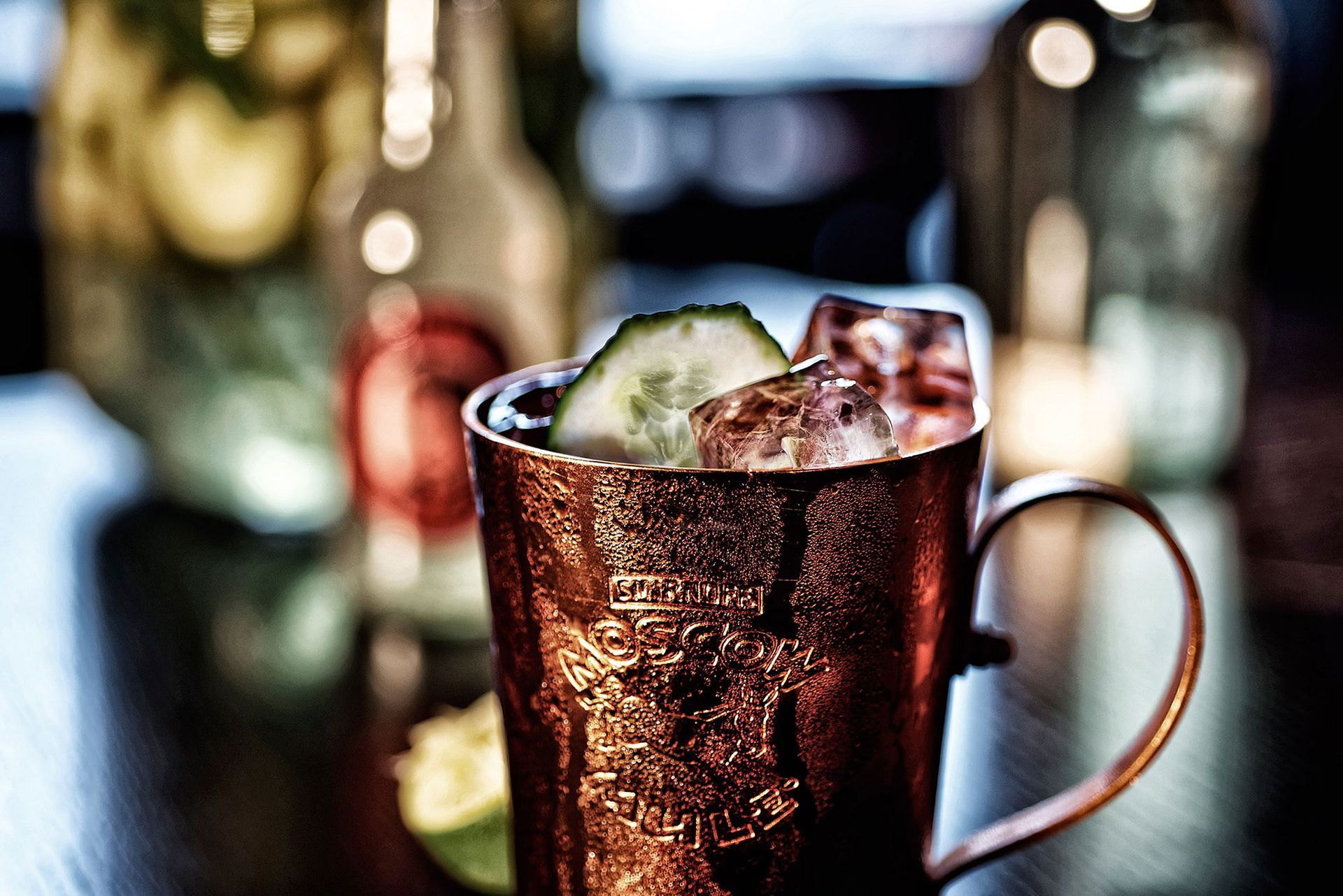 Kupferbecher mit Cocktail, Eis, Gurkenscheibe, Flaschen im Hintergrund.