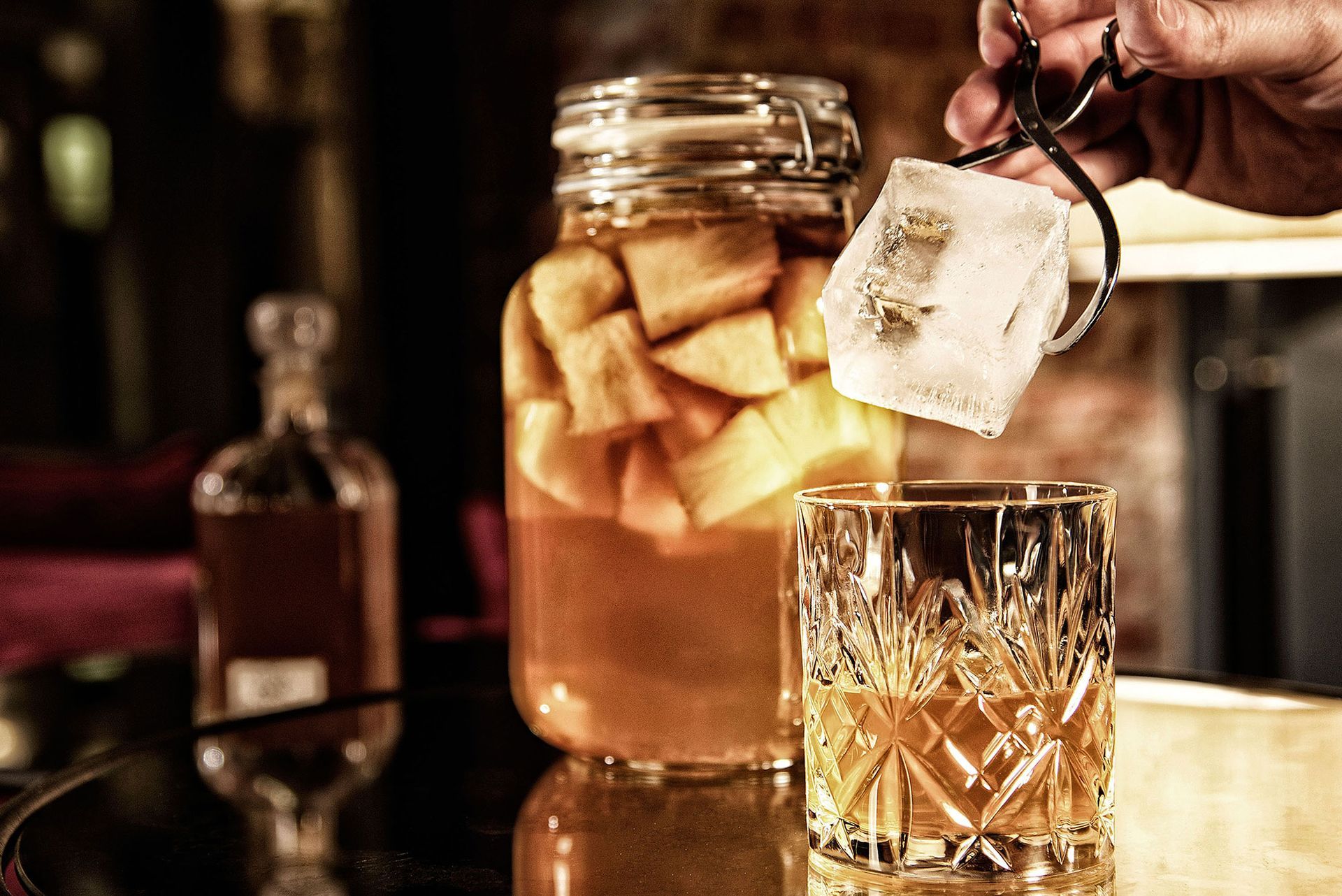 Eine Hand lässt einen Eiswürfel in ein Cocktailglas fallen, daneben ein Einmachglas mit Zutaten und eine Flasche.
