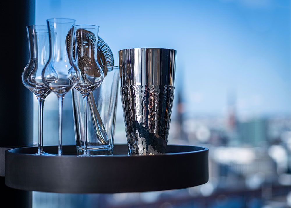 Cocktailshaker, Gläser und Rührlöffel auf einem Tablett vor einem Fenster mit verschwommener Aussicht auf die Stadtlandschaft.