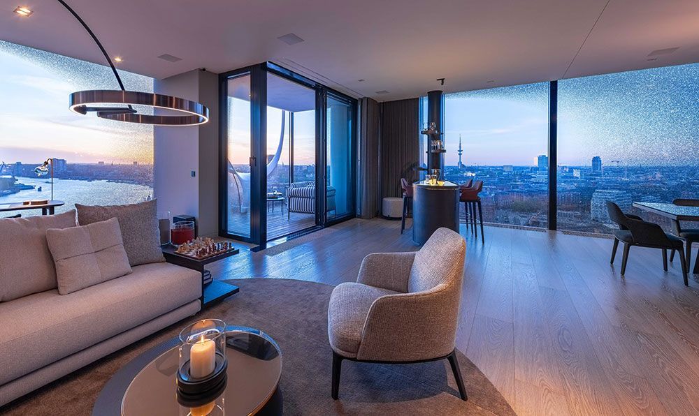 Modernes Wohnzimmer mit Blick auf die Stadt in der Abenddämmerung: beiges Sofa, Sessel, Holzböden, große Fenster, runde Lampe.