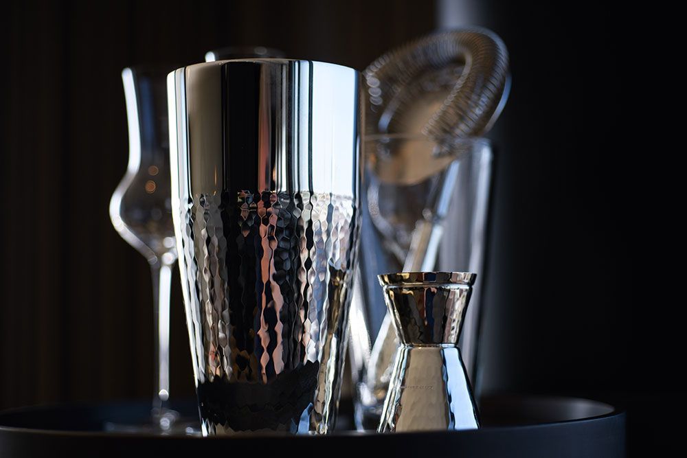 Cocktailshaker, Messbecher und Sieb auf schwarzer Oberfläche vor dunklem Hintergrund, die das Licht reflektieren.