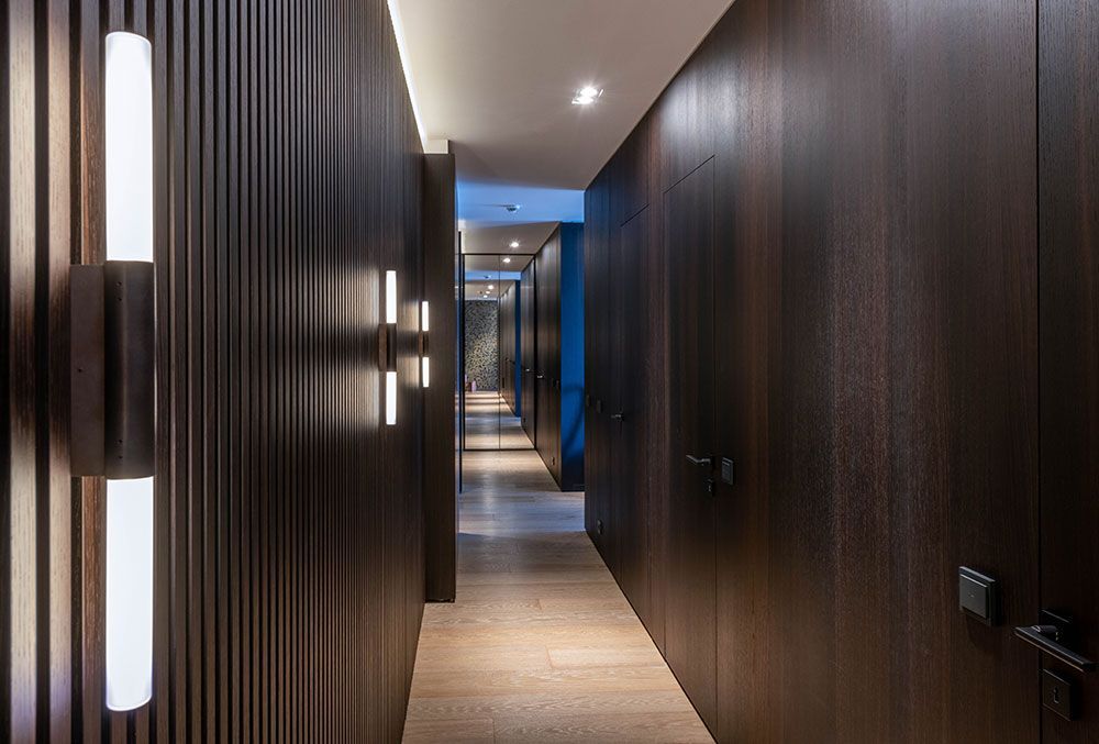 Ein dunkel holzgetäfelter Flur mit modernen Leuchten führt zu einem beleuchteten Türbereich.
