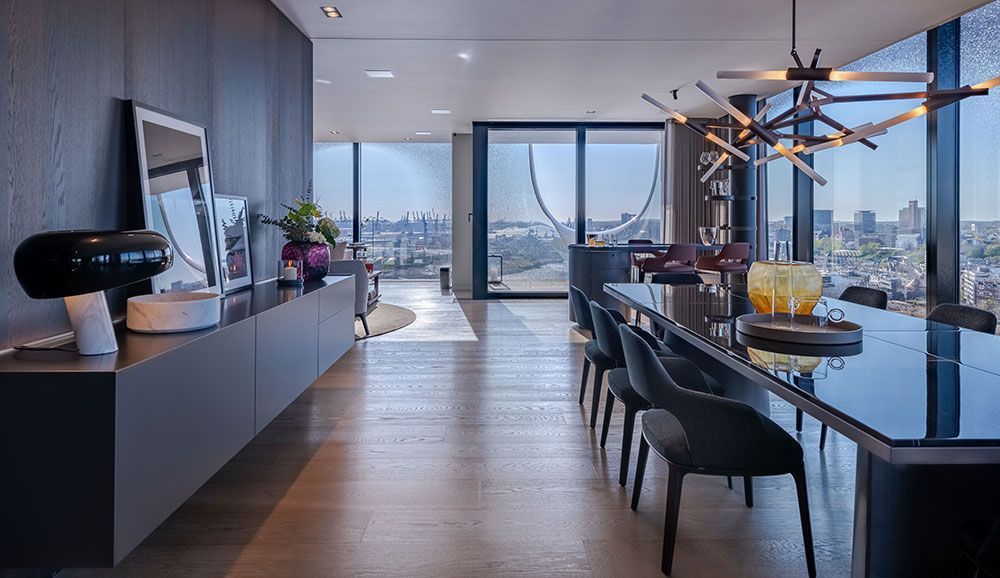Langer Esstisch in moderner Wohnung mit Stadtblick, dunkelgrauen Möbeln und dekorativer Beleuchtung.