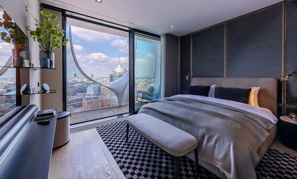 Schlafzimmer mit großen Fenstern, Stadtblick, grau-goldener Dekoration, Bett mit weiß-grauer Bettwäsche.