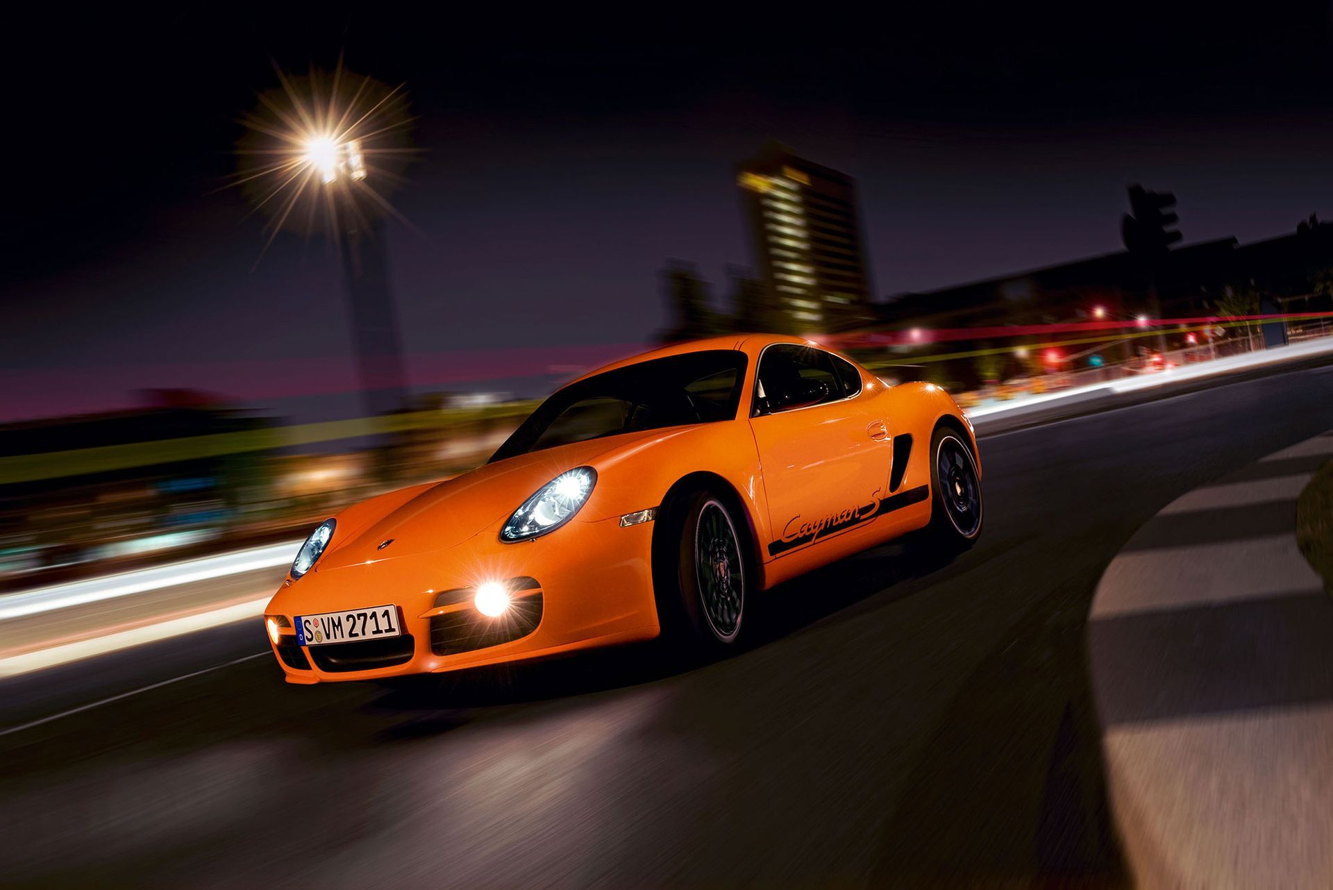 Ein orangefarbener Porsche-Sportwagen fährt nachts auf einer Rennstrecke; im Hintergrund sind verschwommene Lichter und Gebäude zu sehen.