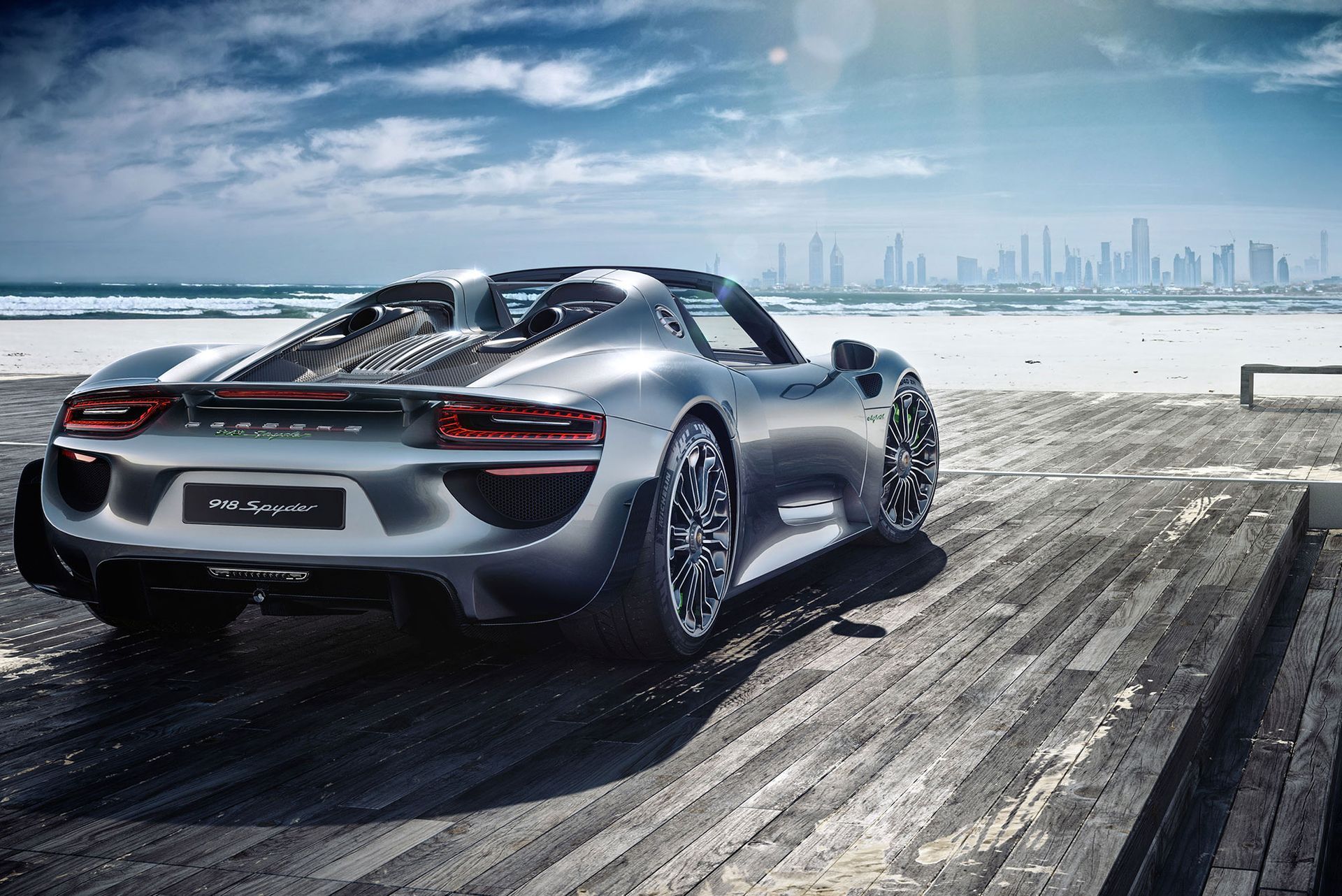 Ein silberner Porsche 918 Spyder parkt auf einem Holzsteg in Strandnähe, im Hintergrund zeichnet sich die Skyline der Stadt unter sonnigem Himmel ab.