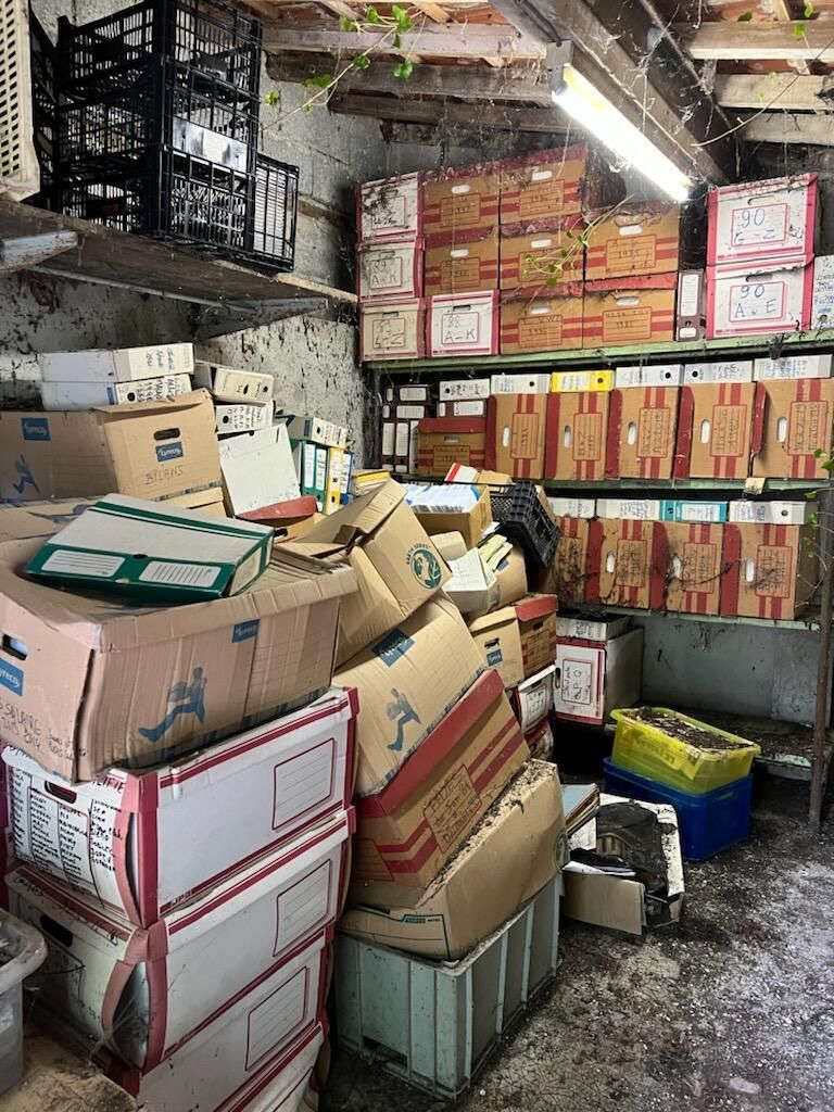 Pièce encombrée : meubles enfouis sous des tas d'ordures et de débris, papiers éparpillés sur le sol.