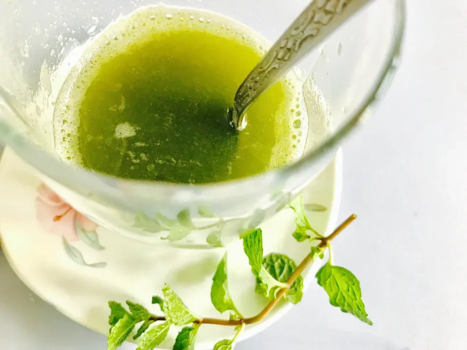 Vaso de té verde con una cuchara y una ramita de menta sobre un fondo blanco.
