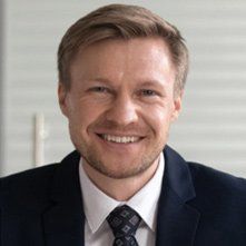 A smiling person with light brown hair wearing a dark blue suit, white collared shirt, and a patterned tie.