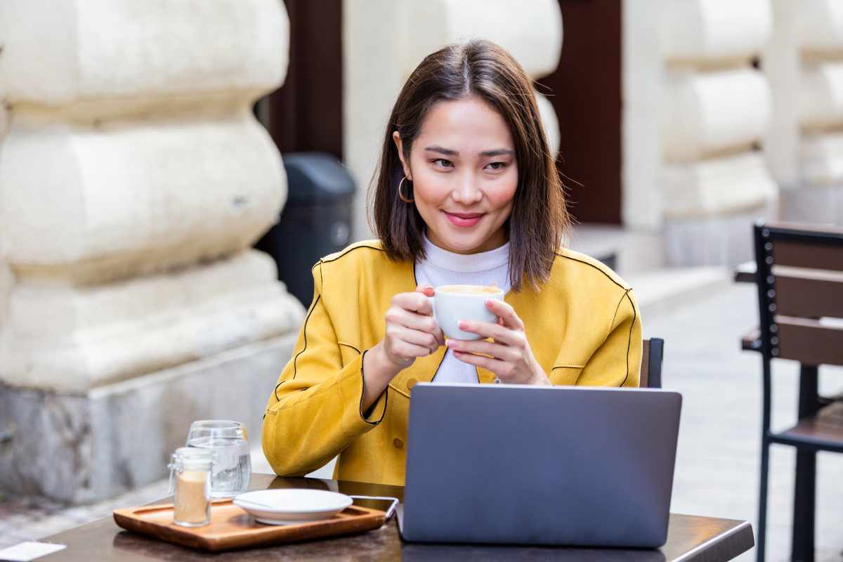 A person in a yellow jacket sits at an outdoor cafe with a laptop, holding a white coffee cup and smiling.