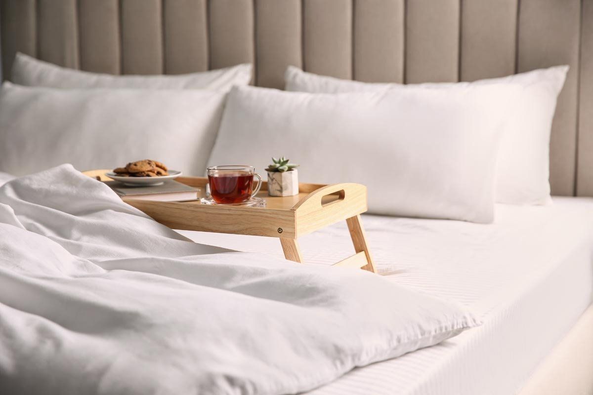 A wooden breakfast tray with a tea cup and cookies sits on a white bed with pillows in a bright bedroom.