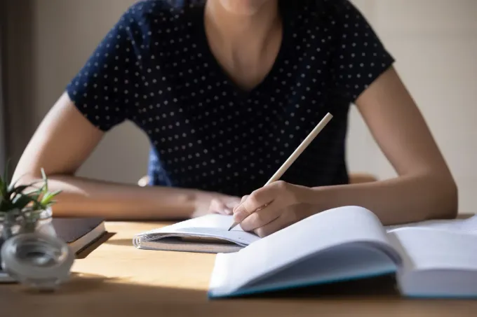 Una persona escribe con lápiz en un cuaderno en un escritorio. Se ven libros y una pequeña planta.