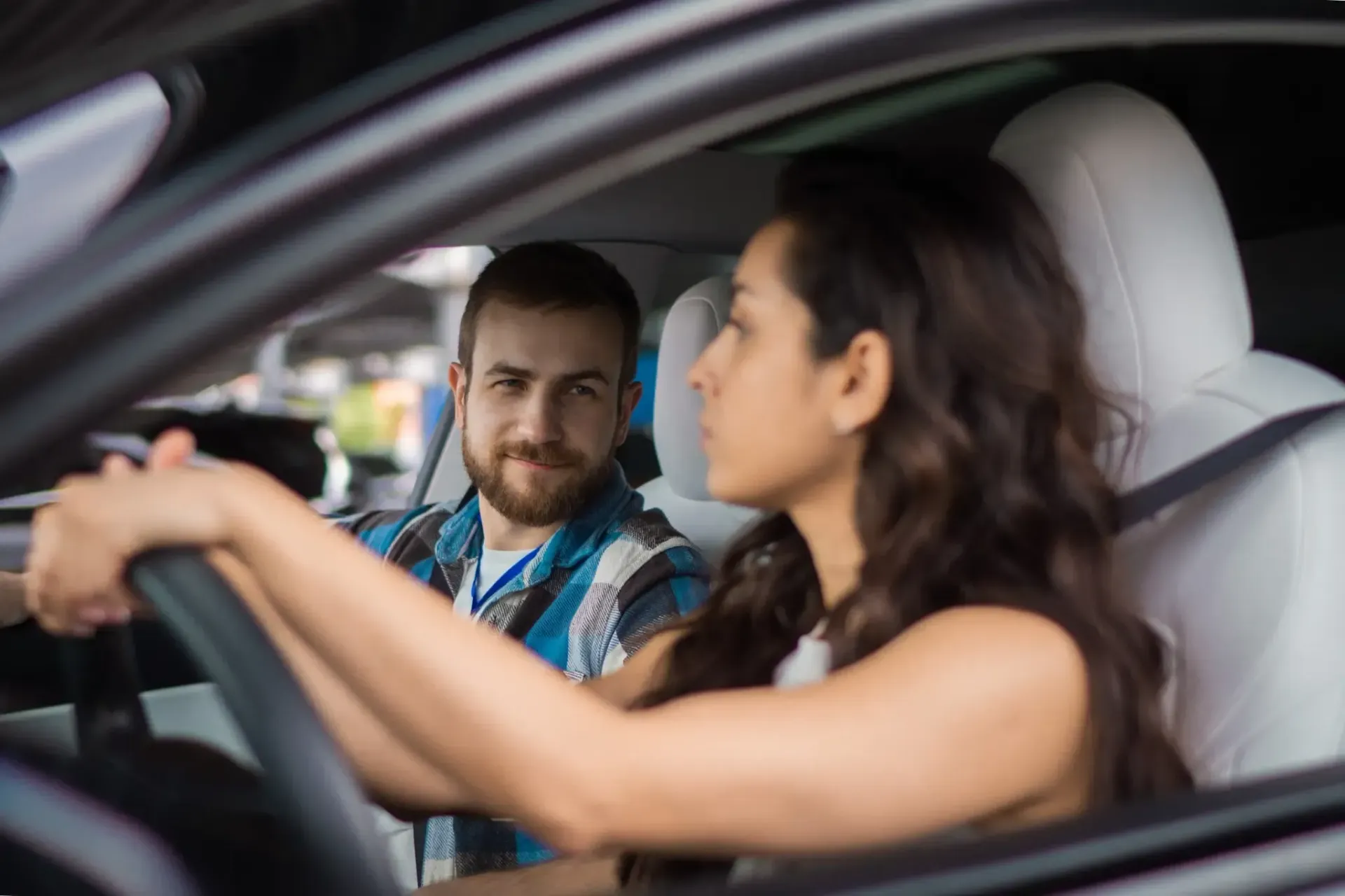 Mujer conduciendo, hombre en el asiento del pasajero mirándola. Interior del coche con asientos blancos.
