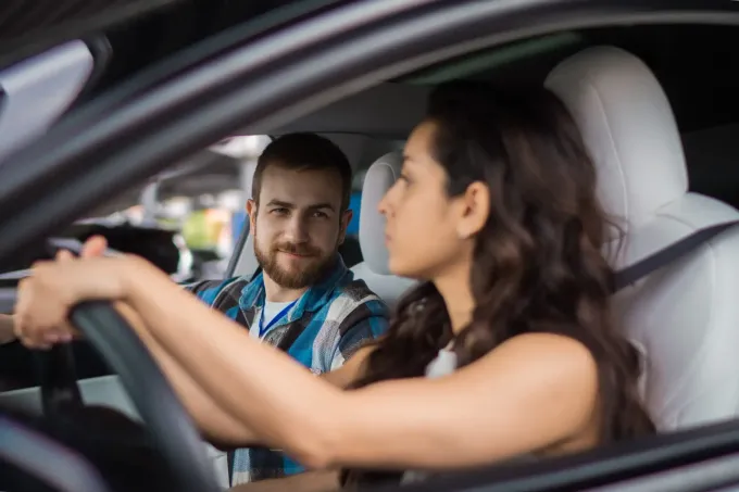 Mujer conduciendo, hombre en el asiento del pasajero mirándola. Interior del coche con asientos blancos.