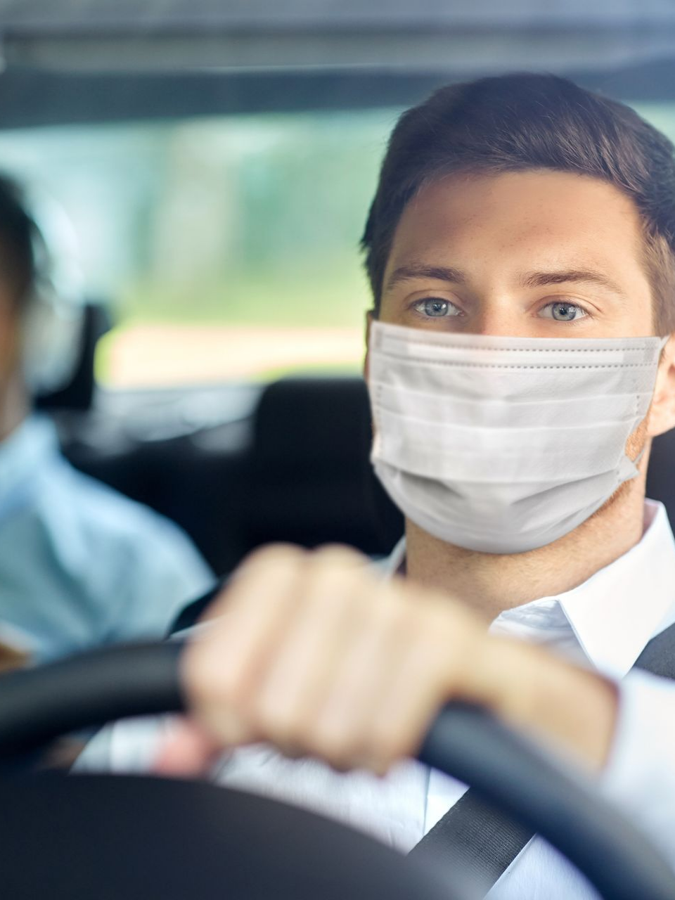 Un homme portant un masque conduit une voiture, concentré sur la route, avec un passager visible.