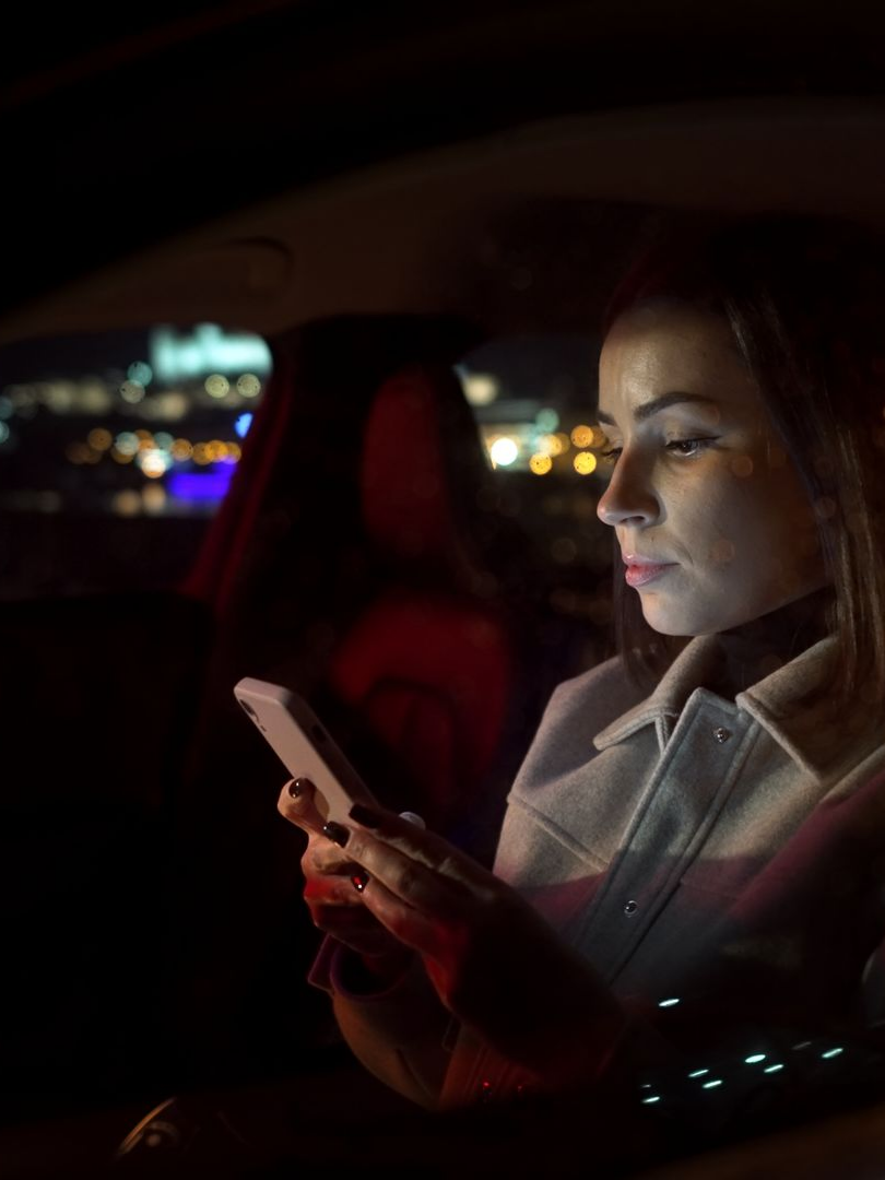 Une femme, assise dans une voiture, regarde son téléphone la nuit.