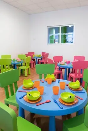 Comedor infantil colorido: mesas redondas azules, sillas verdes y rosas, conjunto con vajilla naranja y verde.