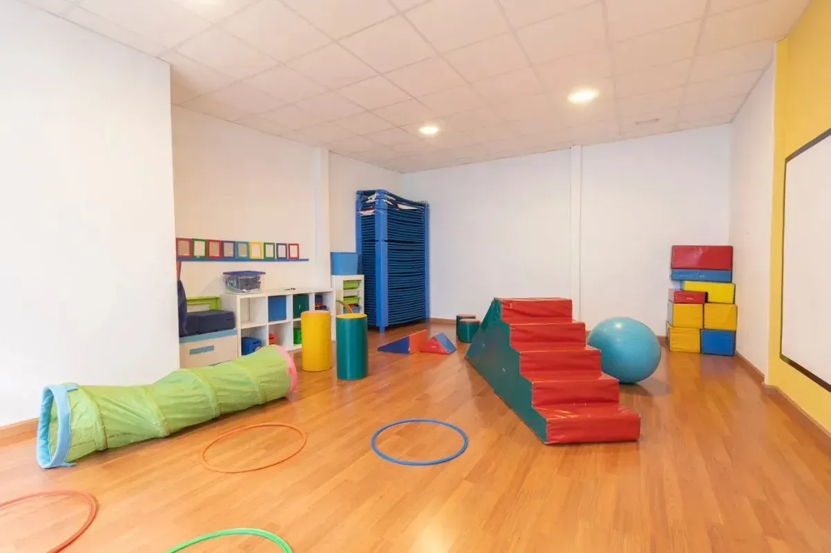 Sala de juegos con suelo de madera, juguetes, túnel, escaleras y una pelota. Paredes blancas con detalles en amarillo.