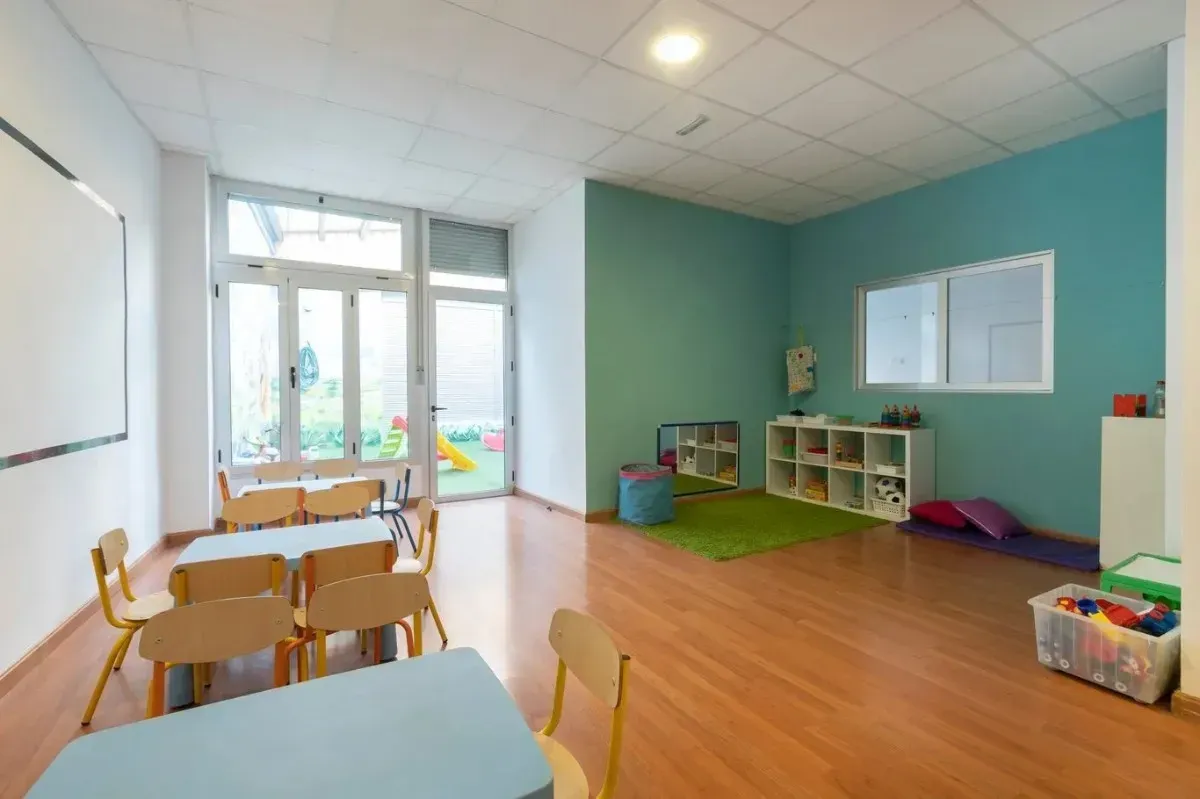 Aula vacía con mesas, sillas y zona de juegos visibles a través de la puerta.