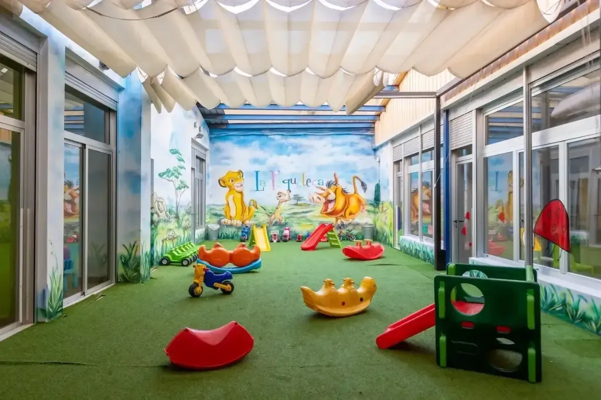 Sala de juegos con césped artificial, juegos infantiles coloridos y un mural.