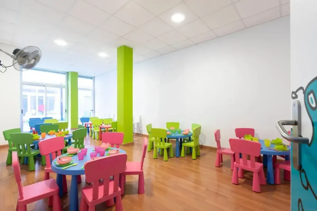 Mesas y sillas infantiles de colores vivos llenan la sala de juegos. Suelo de madera y paredes verdes y blancas.