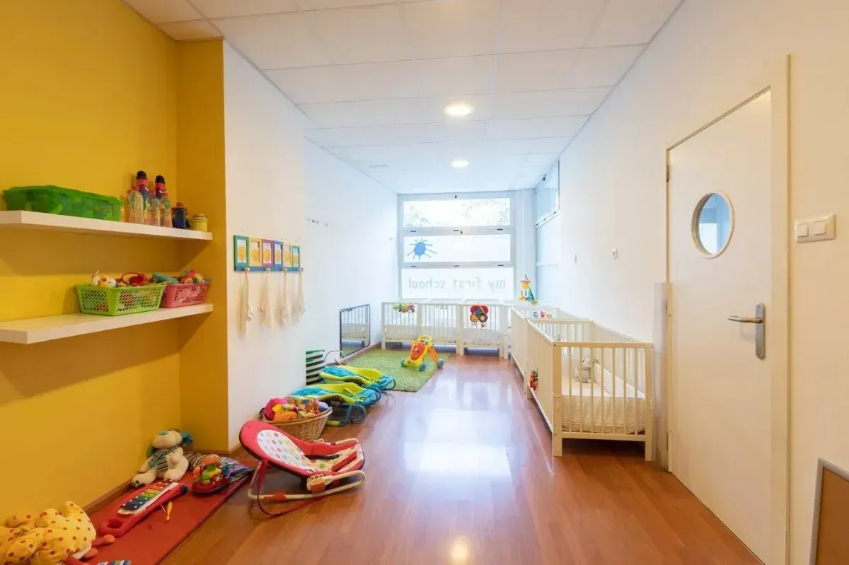 Pasillo de una guardería pintada de colores brillantes, con cunas, juguetes y una ventana.