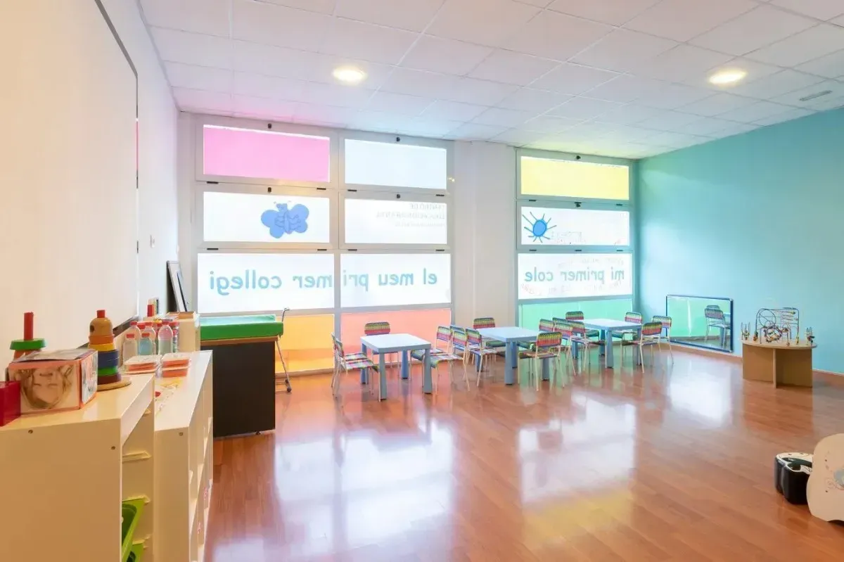 Aula con ventanas coloridas, mesas y sillas, y materiales de arte en un estante.