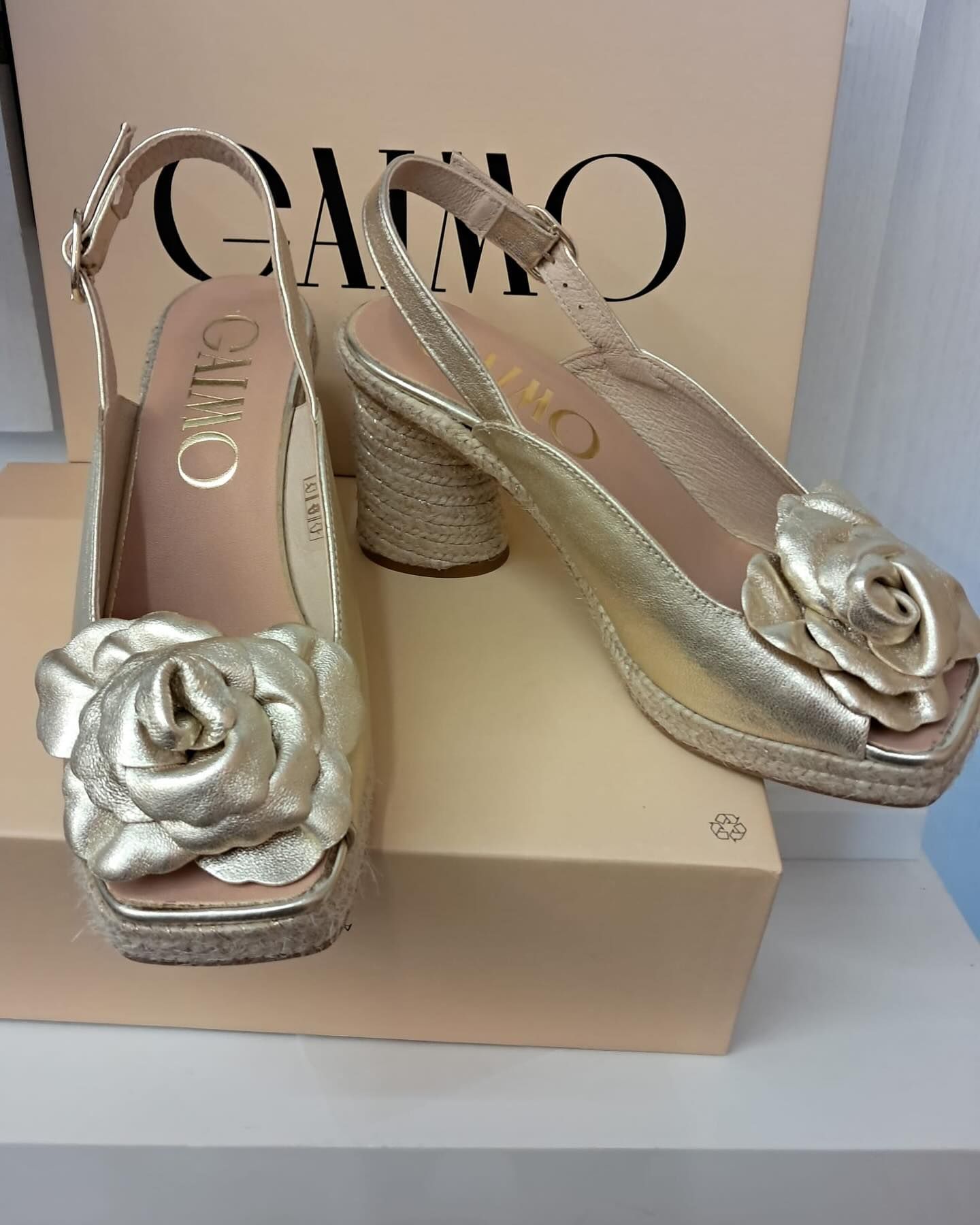 Zapatos de tacón dorados, abiertos y con detalle floral, que reposan sobre una caja con la marca 