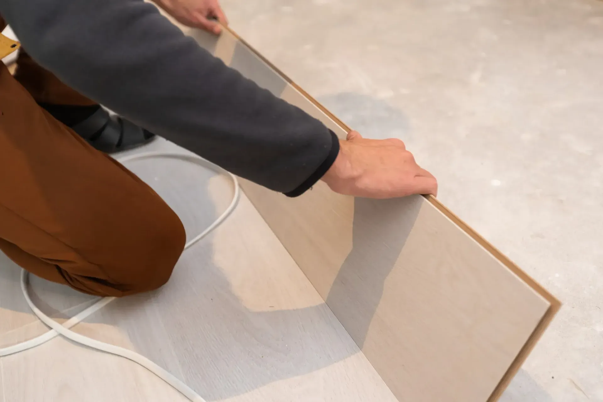 Persona colocando una tabla de madera en el suelo. Manos apoyadas en la tabla, agachado. Tonos blancos y marrones.