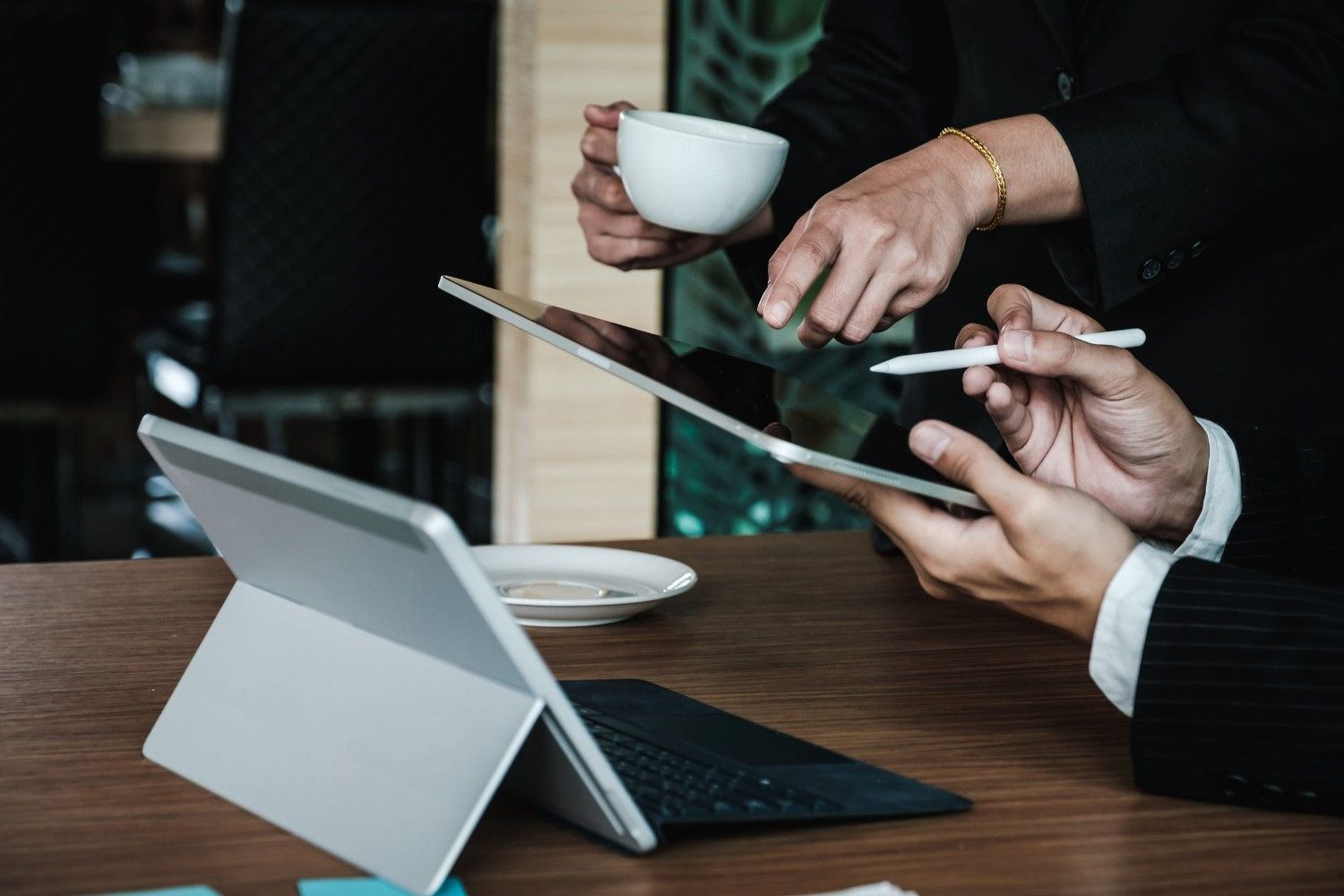 Personas usando una tableta y un portátil en la mesa de una cafetería; una de ellas sostiene una taza de café