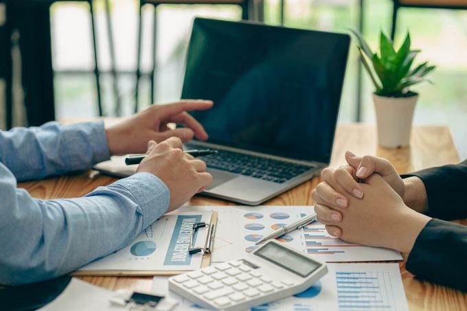 Dos personas hablando de finanzas en un escritorio con un portátil, una calculadora y documentos