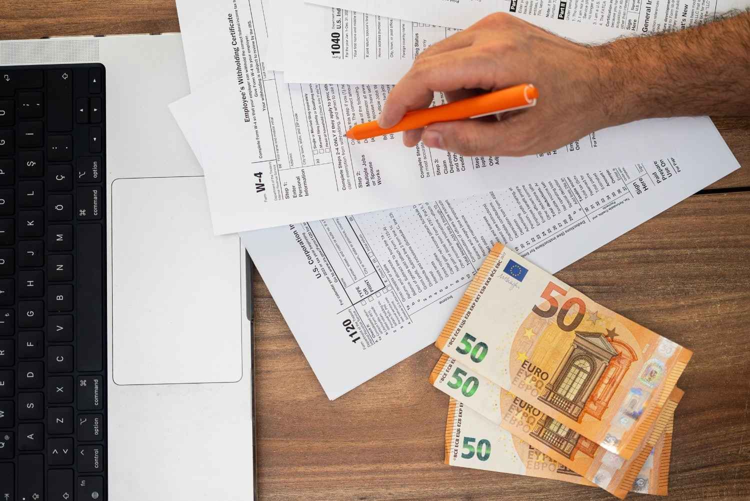 Una mano sostiene un bolígrafo naranja sobre unos documentos en un escritorio, junto a un teclado y unos billetes de euro