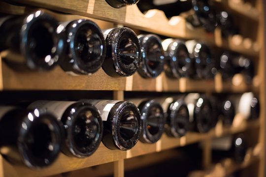 Des rangées de bouteilles de vin noir sont stockées horizontalement sur des étagères en bois dans une cave.