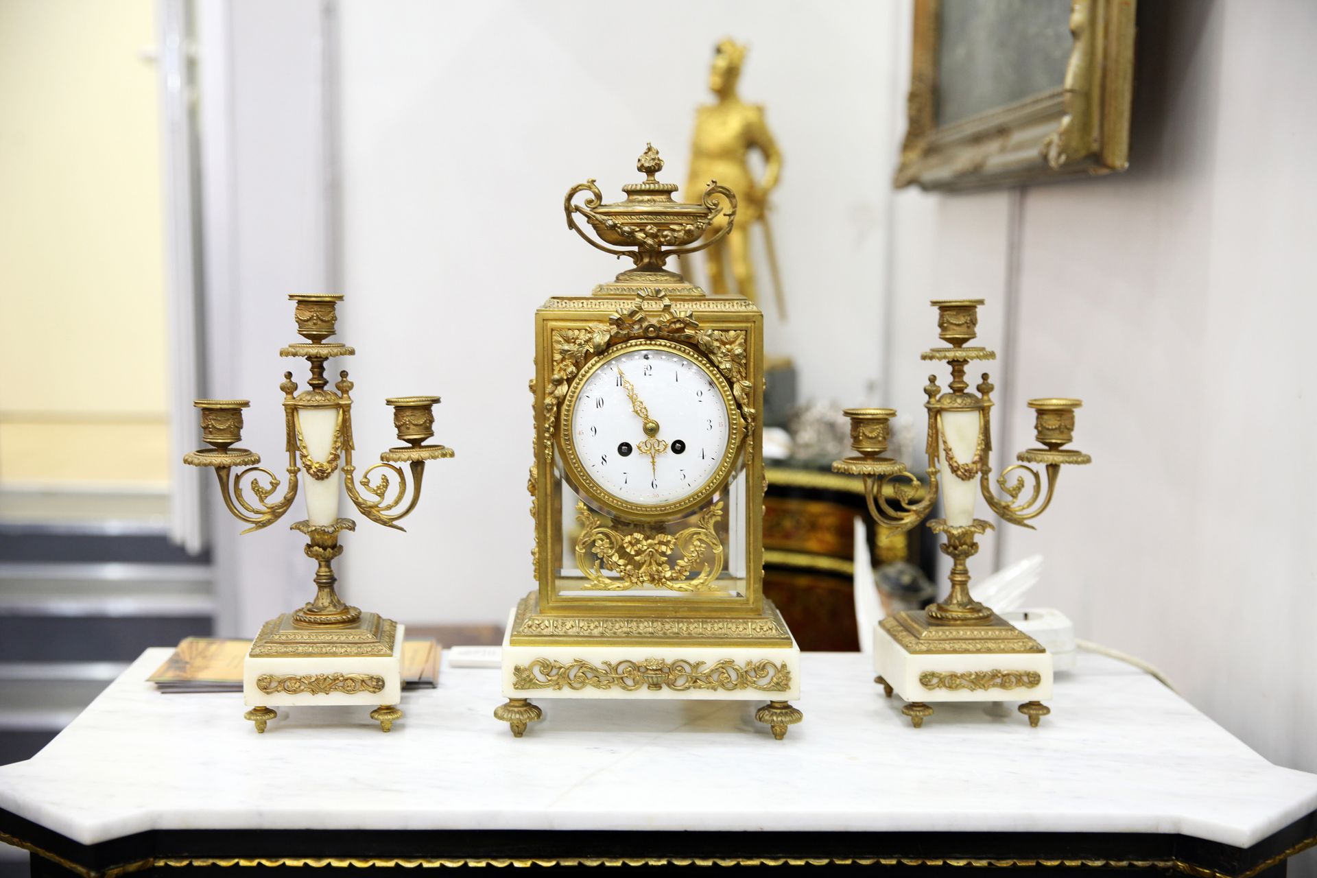 Pendule de cheminée dorée ornée avec candélabres assortis sur une cheminée blanche dans une pièce formelle.