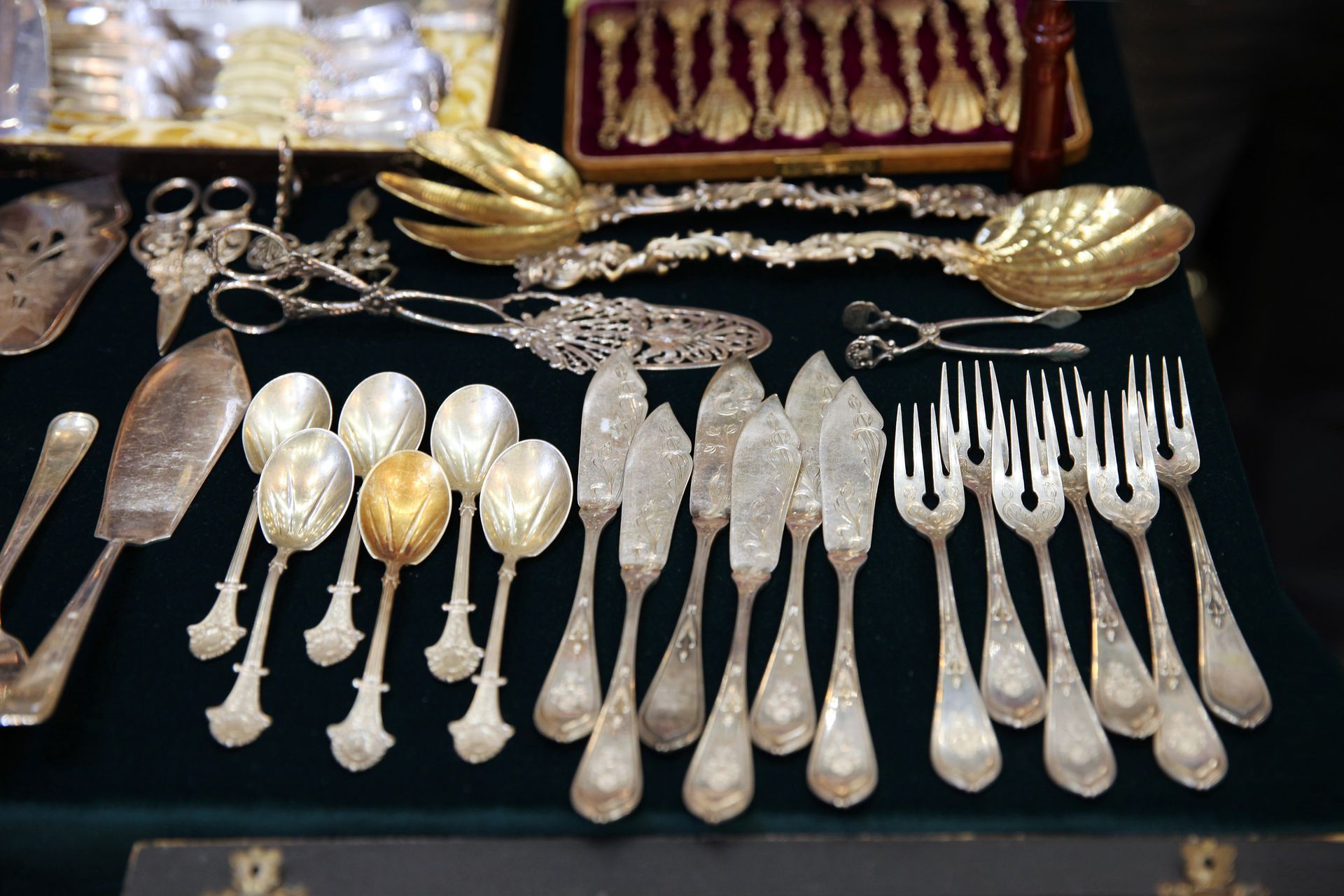 Assortiment de couverts et de cuillères de service en argent et en or finement ouvragés, présentés sur un fond en tissu noir.