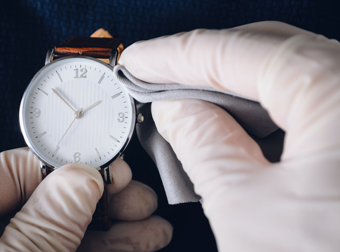 Des mains gantées nettoient une montre-bracelet en argent avec un chiffon sur fond sombre.