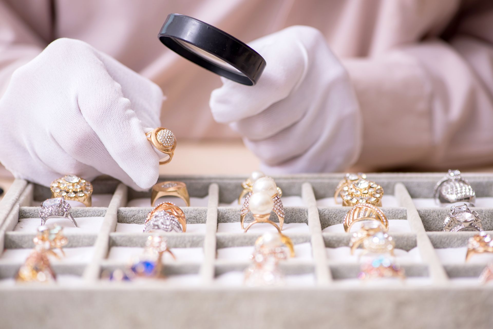 Un bijoutier en gants blancs examine des bagues exposées sur un plateau à l'aide d'une loupe grossissante.