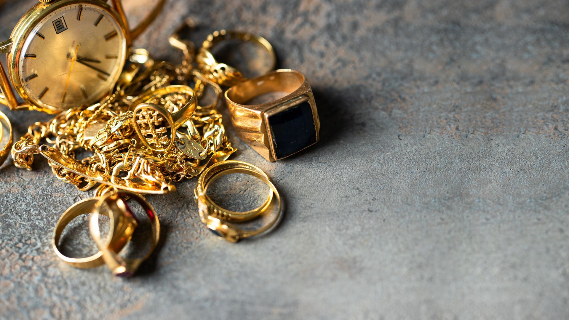 Des bijoux en or et une montre-bracelet éparpillés sur une surface grise, avec deux bagues au premier plan.