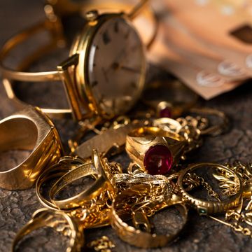 Des bagues en or et des bijoux empilés sur une surface sombre, avec une montre de poche en arrière-plan.