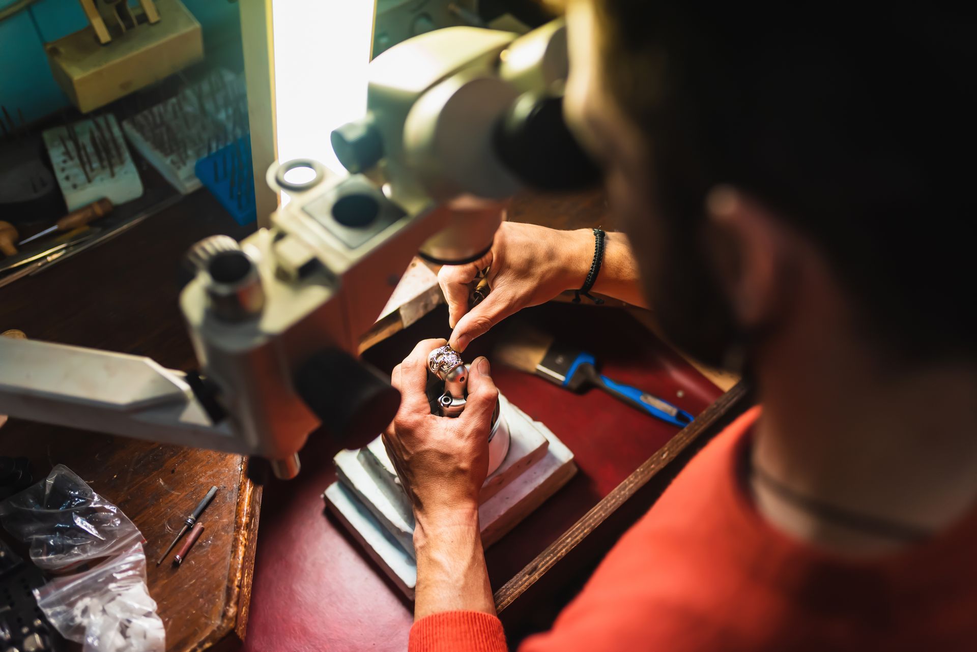 Une personne utilise un microscope sur un établi tout en manipulant un petit objet sous une lampe de bureau.