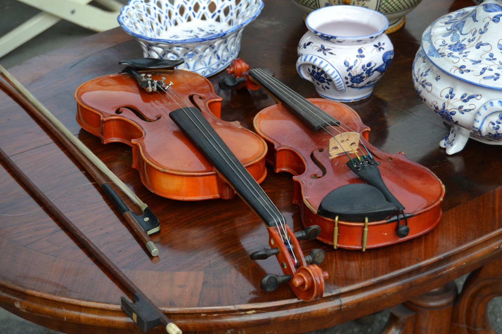 Trois violons et un archet sur une table en bois, à côté de bols et de tasses en porcelaine bleue et blanche.