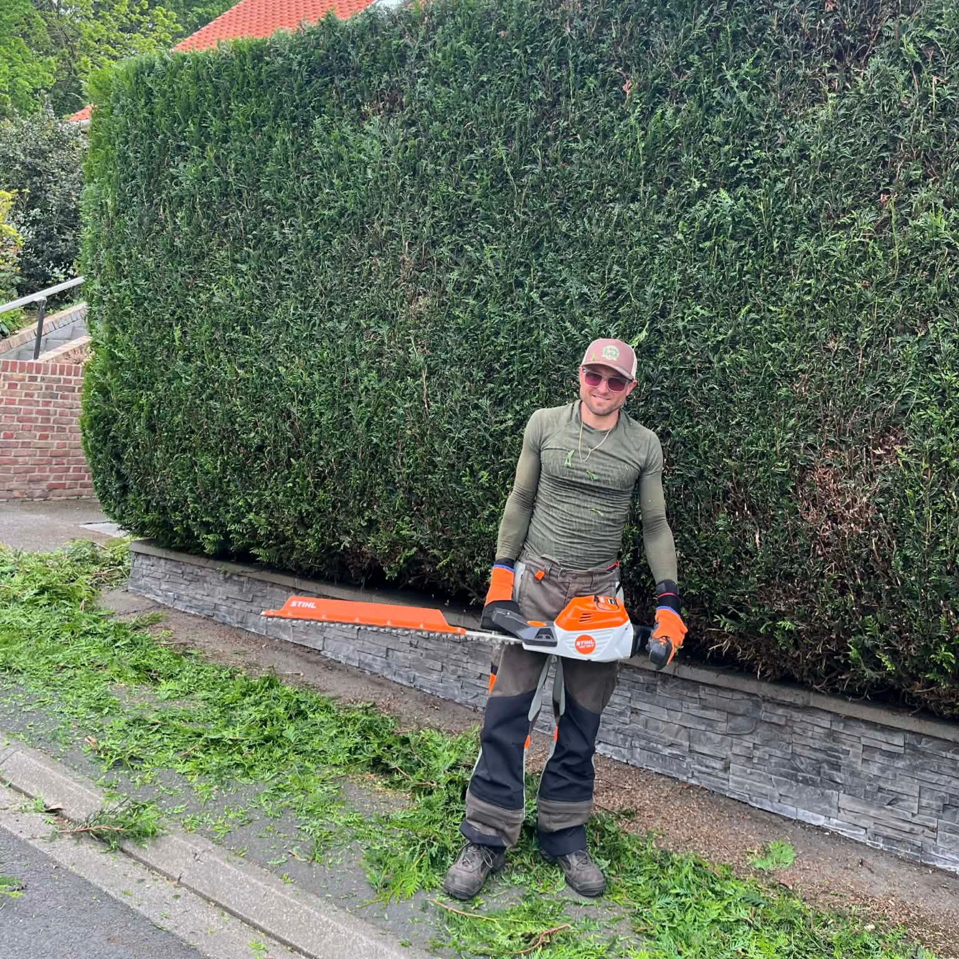 Une personne en tenue de travail et gants orange se tient à côté d'une grande haie, tenant un taille-haie sur un trottoir.