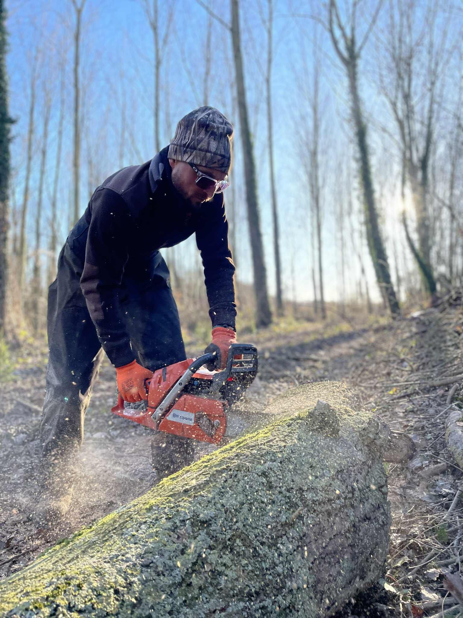 Une personne utilise une tronçonneuse pour couper un arbre tombé à l'extérieur, des copeaux de bois volent dans tous les sens.