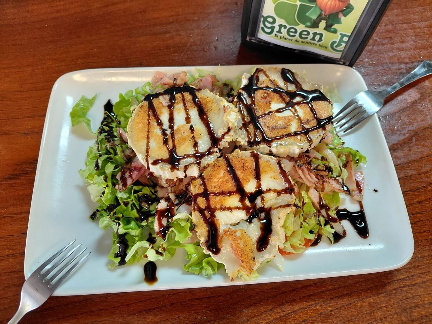 Ensalada con pollo a la parrilla y un chorrito de vinagre balsámico servida en un plato blanco, con tenedores sobre una mesa de madera.