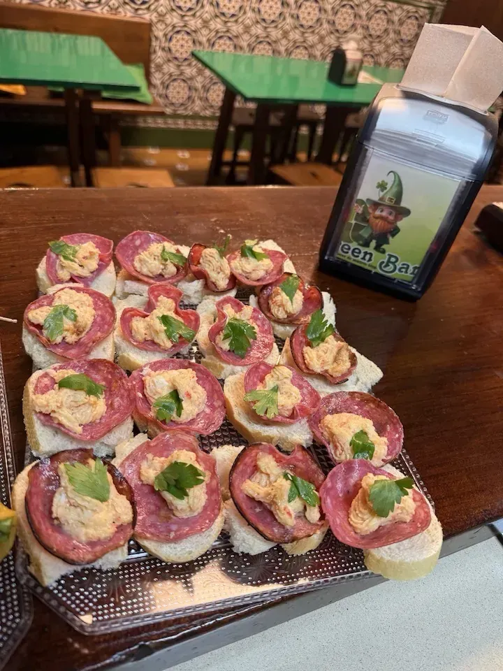Aperitivos en bandeja: salami, crema para untar, cilantro y pan tostado. En un ambiente de bar.