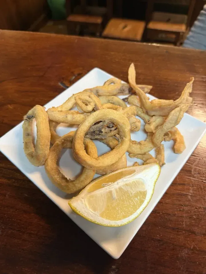 Anillos y tentáculos de calamares fritos en un plato blanco con rodaja de limón, sobre una barra de madera.