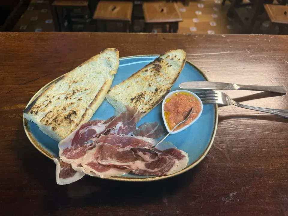 Pan tostado, prosciutto y salsa de tomate en un plato azul, con cubiertos sobre una mesa de madera.