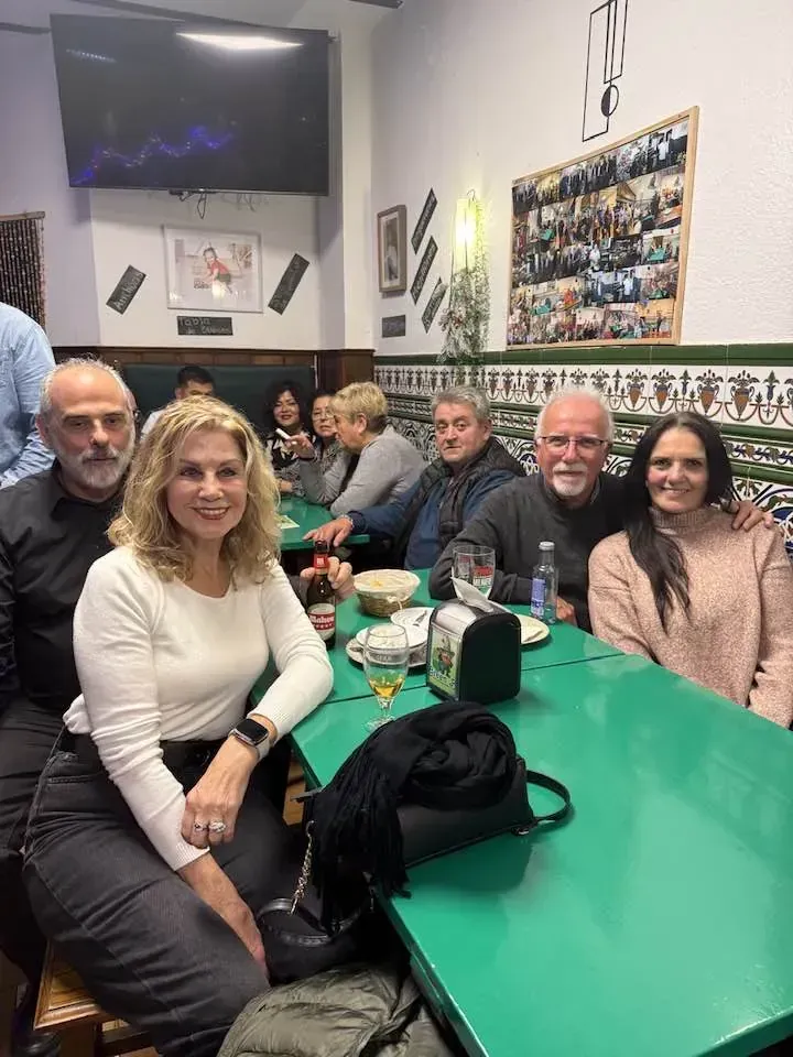 Un grupo de personas sentadas en una mesa verde en un restaurante. Algunas sonríen. Hay un televisor colgado en la pared.