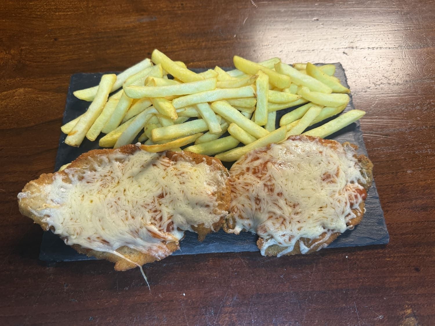 Plato gratinado con queso y patatas fritas, servido en una tabla oscura sobre una mesa de madera.